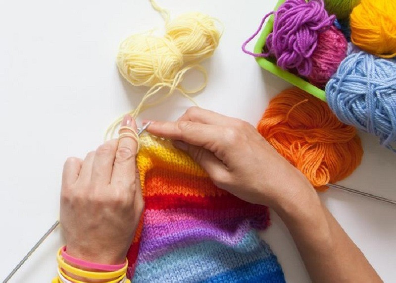 A top down shot of two hands knitting a rainbow stripe piece of fabric